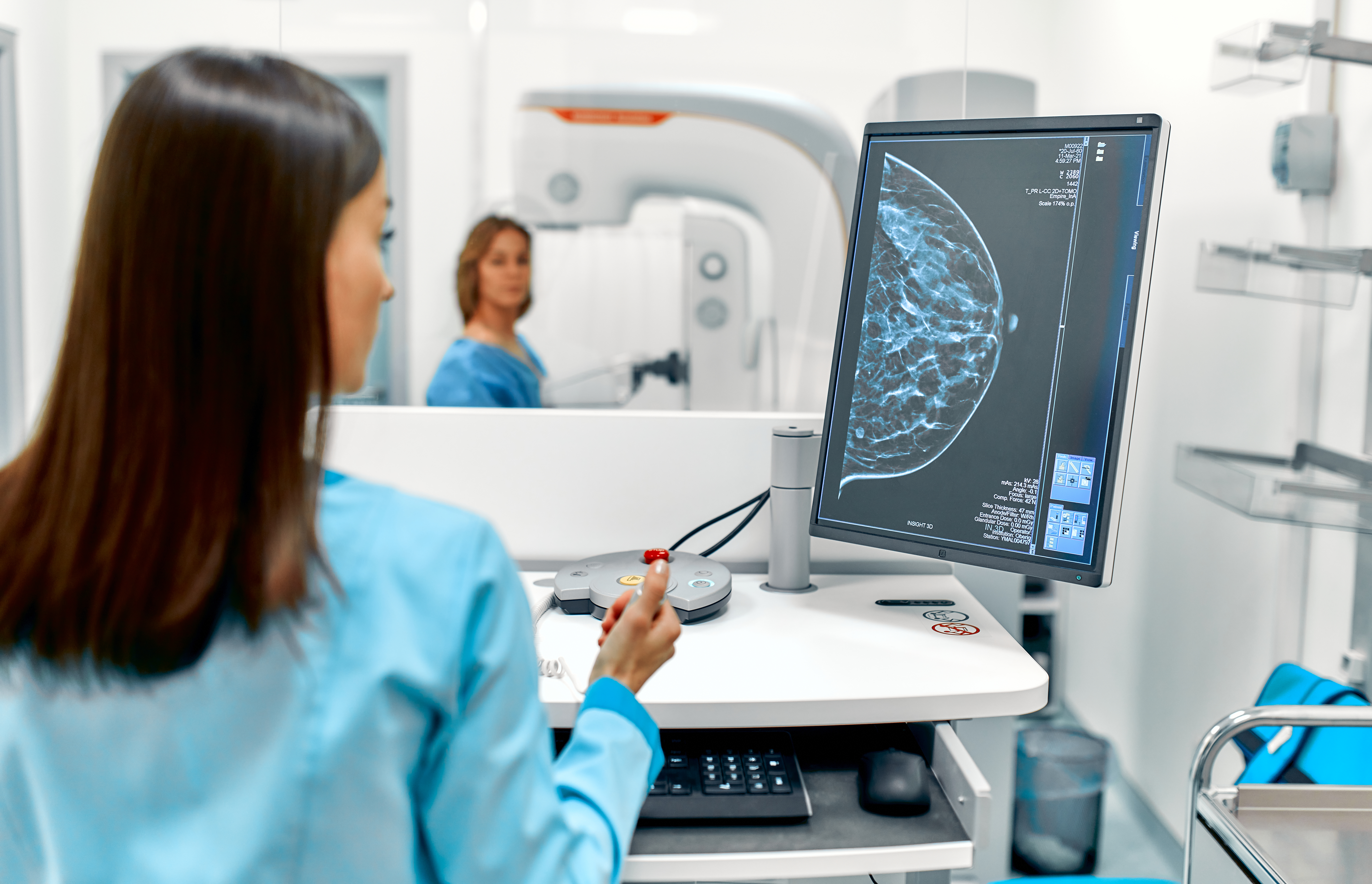 A healthcare professional in scrubs reviews a mammogram image on a computer monitor while another clinician prepares a mammography machine in a clinical imaging room.